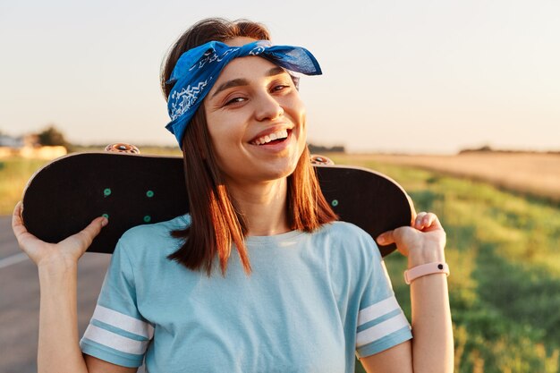 Außenaufnahme einer zufriedenen, fröhlichen Frau mit dunklem Haar, die Skateboard über den Schultern hält und mit einem zahnigen Lächeln direkt in die Kamera schaut und im Sommer Skateboarding genießt.