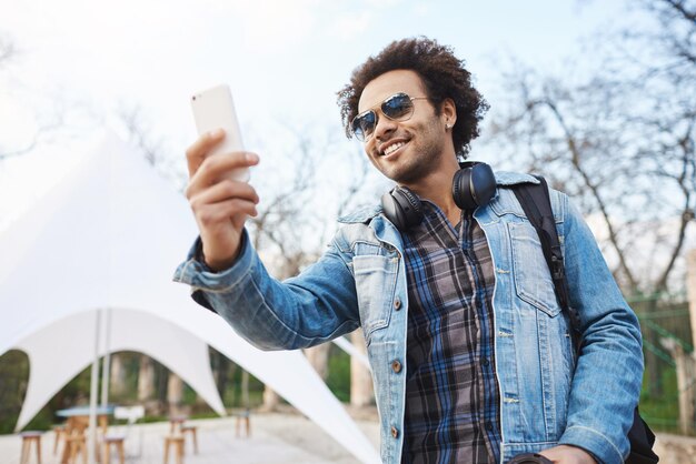 Außenaufnahme eines attraktiven jungen Afroamerikaners mit Afro-Frisur und Kopfhörern über dem Hals, der trendige Kleidung und Brille trägt und ein Smartphone hält, während er eine Straßenband im Park aufnimmt