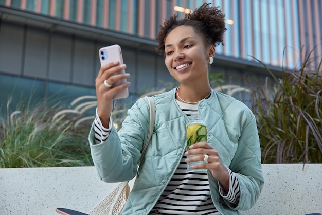 Außenaufnahme eines glücklichen Teenager-Mädchens mit zwei lockigen Brötchen macht ein Selfie über Smartphone-Getränke Detox-Getränk trägt einen lässigen gestreiften Pullover und eine Jacke trägt eine Netztasche und verbringt Freizeit in der städtischen Umgebung