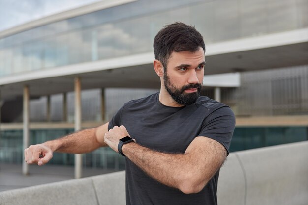 Außenaufnahme eines motivierten bärtigen Mannes, der sich die Arme wärmt, bevor das Training ein lässiges schwarzes T-Shirt von Smartwacth trägt, das in Fernposen gegen Unschärfe konzentriert ist. Sportkonzept.