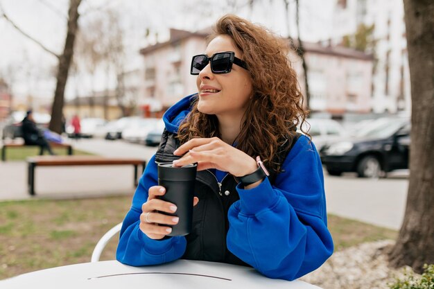 Außenfoto einer attraktiven, lockigen Frau mit Sonnenbrille, die einen hellblauen Pullover und eine Jacke trägt, trinkt an warmen Frühlingstagen Kaffee zum Mitnehmen auf der Sommerterrasse