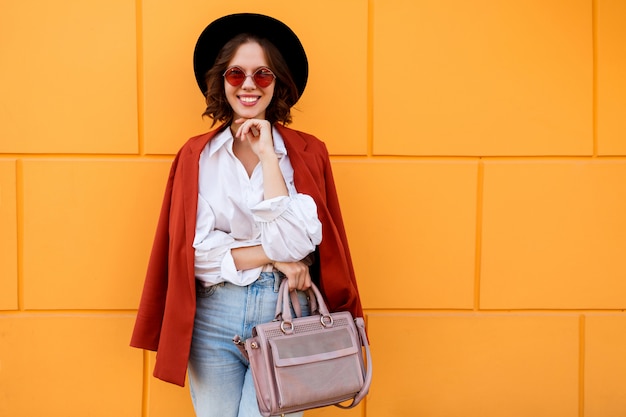 Außennahaufnahmeporträt der glückseligen brünetten kurzhaarigen Frau, die über gelber Wand aufwirft. Trendiger Hut, rosa Brille, weiße Bluse und Jeans.