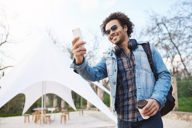 Außenportarit des glücklichen aufrichtigen Afroamerikanermannes in der stilvollen Brille und im Jeansmantel, die Tasse Kaffee halten und Foto auf Smartphone machen, während breit während des Spaziergangs im Park lächeln.