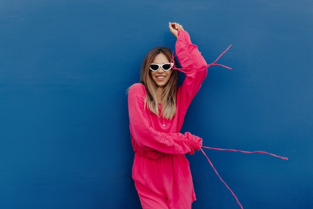 Außenporträt der attraktiven stilvollen jungen Frau gekleidet rosa Kleid und weiße Sonnenbrille, die über isolierte blaue Wand aufwirft