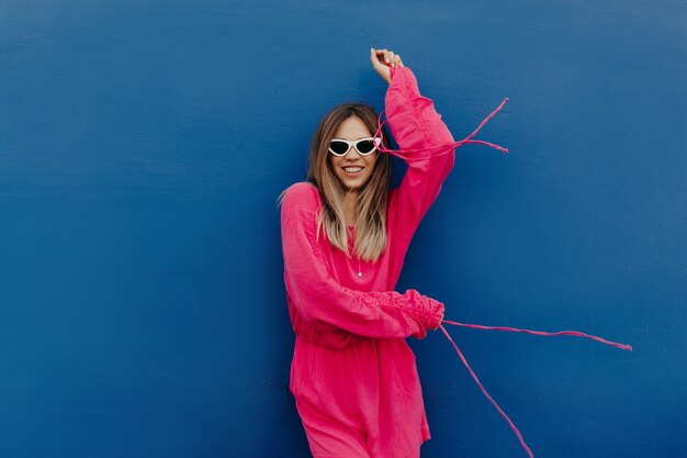 Außenporträt der attraktiven stilvollen jungen Frau gekleidet rosa Kleid und weiße Sonnenbrille, die über isolierte blaue Wand aufwirft