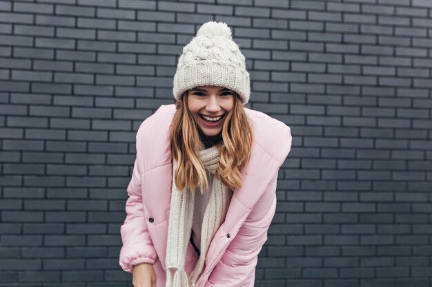 Kostenloses Foto außenporträt der blithesome frau im rosa mantel. attraktives blondes mädchen im winterhut, der vor ziegelmauer aufwirft.
