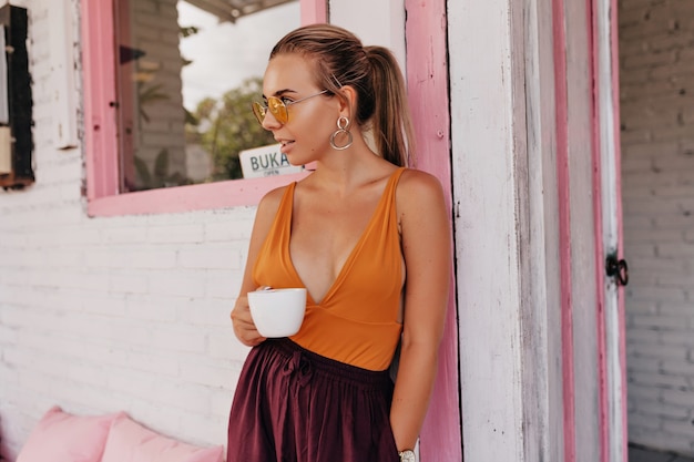 Außenporträt der charmanten Dame im orangefarbenen T-Shirt und in den runden Gläsern, die eine Tasse Kaffee halten