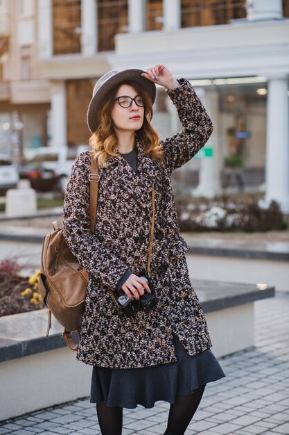 Außenporträt der eleganten jungen Dame mit braunem Rucksack, der Mantel und Hut trägt. Attraktive Frau mit lockigem Haar, die am Telefon spricht, während sie Kaffee auf der Straße trinkt und Freunde wartet.