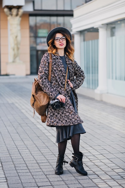 Kostenloses Foto außenporträt der eleganten jungen dame mit braunem rucksack, der mantel und hut trägt. attraktive frau mit lockigem haar, die am telefon spricht, während sie kaffee auf der straße trinkt und freunde wartet.