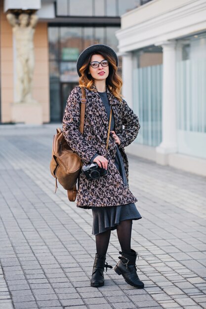 Außenporträt der eleganten jungen Dame mit braunem Rucksack, der Mantel und Hut trägt. Attraktive Frau mit lockigem Haar, die am Telefon spricht, während sie Kaffee auf der Straße trinkt und Freunde wartet.