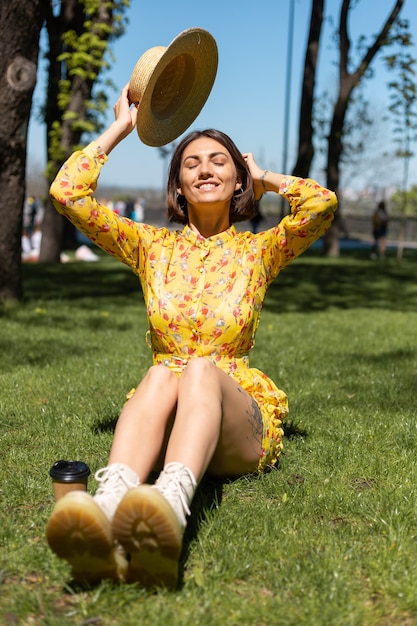 Außenporträt der Frau im gelben Sommerkleid und im Hut, die auf Gras im Park sitzen