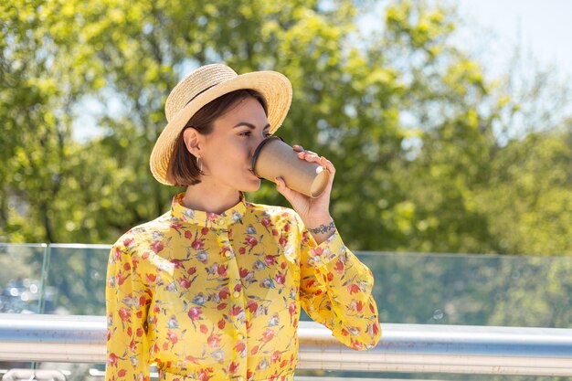 Außenporträt der Frau im gelben Sommerkleid und im Hut mit der Tasse Kaffee, die Sonne genießt