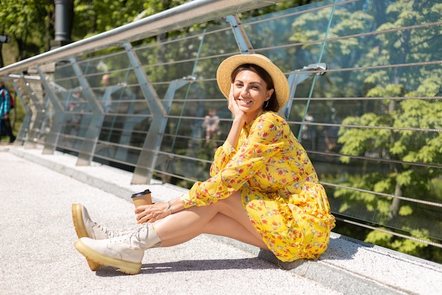 Außenporträt der Frau im gelben Sommerkleid und im Hut mit der Tasse Kaffee, die Sonne genießt