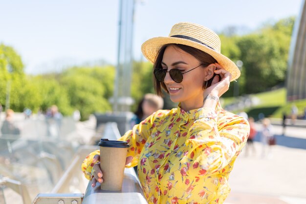 Außenporträt der Frau im gelben Sommerkleid und im Hut mit Tasse Kaffee, die Sonne genießt, steht auf Brücke mit Stadt erstaunliche Ansicht