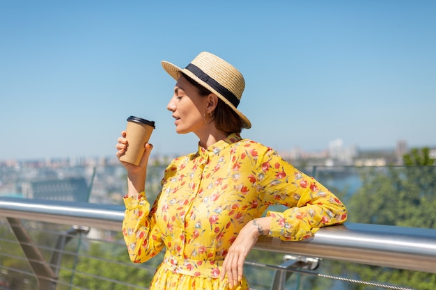 Außenporträt der Frau im gelben Sommerkleid und im Hut mit Tasse Kaffee, die Sonne genießt, steht auf Brücke mit Stadt erstaunliche Ansicht