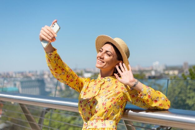 Außenporträt der Frau im gelben Sommerkleid und im Hut nehmen Selfie am Telefon, steht auf Brücke mit Stadt erstaunliche Ansicht