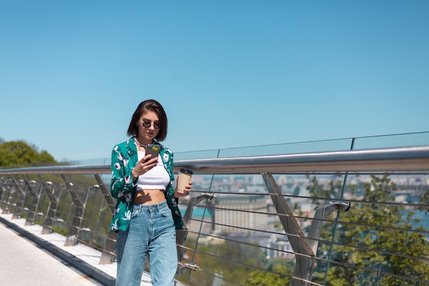 Außenporträt der Frau im lässigen grünen Hemd und in den Jeans am sonnigen Tag geht auf Brücke mit Tasse Kaffee, der auf Telefonbildschirm schaut