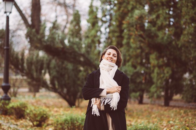 Außenporträt der Frau im Park, der schwarzen Wintermantel und weißen Schal trägt