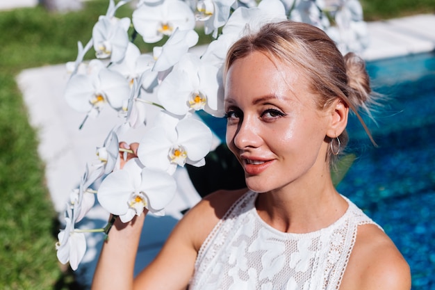 Außenporträt der Frau im weißen Hochzeitskleid, das nahe dem blauen Schwimmbad mit Blumen sitzt