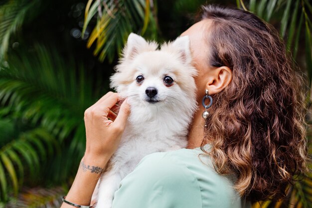 Außenporträt der gelockten europäischen gebräunten Frau hält glücklichen pomeranischen Spitzhund