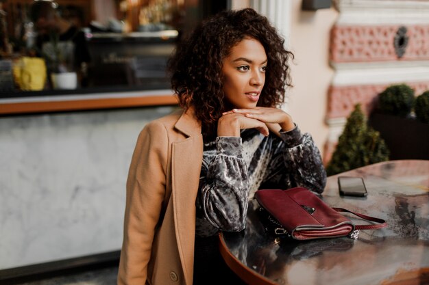 Außenporträt der schönen lächelnden schwarzen Frau mit stilvollen Afro-Haaren, die im Café in Paris sitzen.