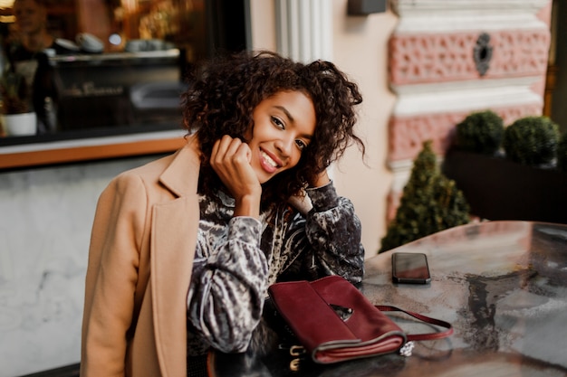 Außenporträt der schönen lächelnden schwarzen Frau mit stilvollen Afro-Haaren, die im Café in Paris sitzen.