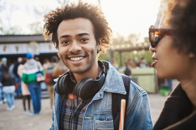 Außenporträt des charmanten afroamerikanischen Mannes, der mit Freund im Park geht und Jeanskleidung und Kopfhörer über Hals trägt,