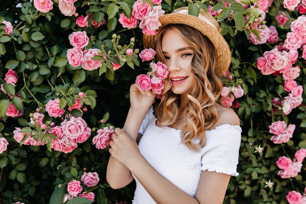 Außenporträt des glücklichen weißen Mädchens, das auf Natur aufwirft. Foto der entspannten Dame mit dem gewellten Haar, das nahe schönem Rosenbusch steht.