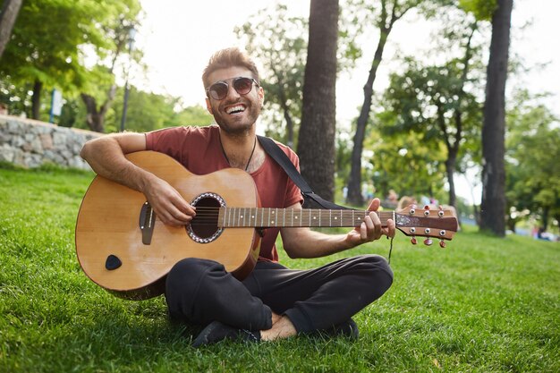 Außenporträt des hübschen Hipster-Mannes, der auf Gras im Park sitzt und Gitarre spielt