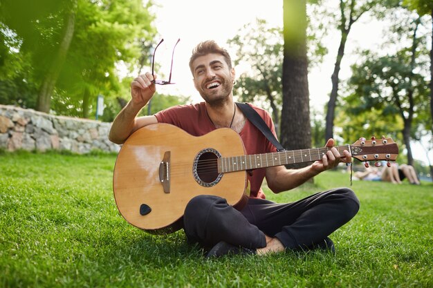 Außenporträt des hübschen Hipster-Mannes, der auf Gras im Park sitzt und Gitarre spielt