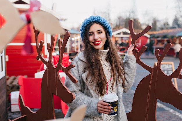 Außenporträt des langhaarigen Mädchens mit der Tasse Kaffee, die nahe Spielzeughirsche im Winterurlaub aufwirft. Foto der reizenden Frau im blauen Hut, der neben Weihnachtsdekoration im Park steht.