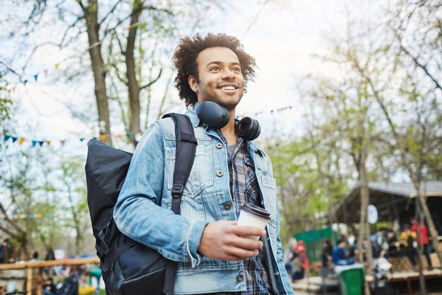 Außenporträt des modischen afroamerikanischen Mannes mit Afro-Haarschnitt, Jeansmantel und Rucksack tragend, während Kaffee halten und beiseite schauen, im Park gehen oder auf jemanden warten.