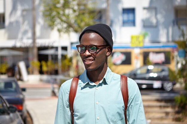 Außenporträt des modischen jungen afroamerikanischen Studenten in der stilvollen Kleidung, die in der städtischen Umgebung geht, sonnigen Morgen genießt, während zu Fuß zum College geht, mit fröhlichem Ausdruck auf seinem Gesicht