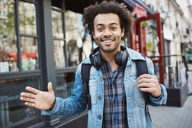 Außenporträt des positiven Afroamerikaners mit der Afro-Frisur, die beim Gehen in der Straße winkt und lächelt, im trendigen Outfit und in den Kopfhörern über Hals trägt.