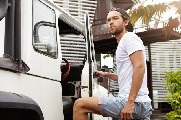 Außenporträt des stilvollen bärtigen Mannes in den Jeansshorts, die in sein weißes Sport Utility Vehicle einsteigen und seine Hand am Griff halten