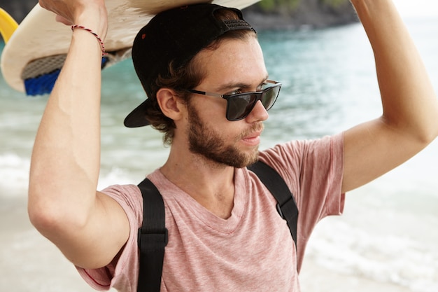 Außenporträt des stilvollen jungen Surfers in der Sonnenbrille, die Surfbrett auf seinem Kopf hält und blaues Meer mit selbstbewusstem und entschlossenem Ausdruck betrachtet