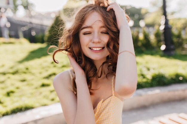 Außenporträt des weißen siegreichen Mädchens mit dem roten lockigen Haar, das auf Natur aufwirft. Lächelnde herrliche Ingwerfrau, die mit geschlossenen Augen im Park steht.