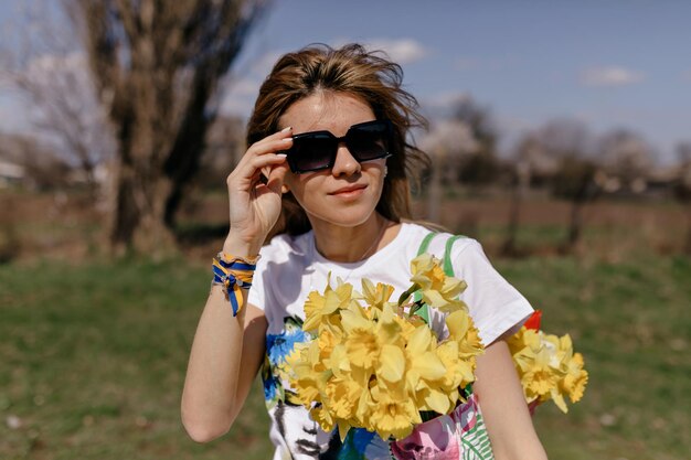 Außenporträt einer stilvollen ukrainischen Frau mit fliegendem Haar, die eine Sonnenbrille trägt und eine Tasche mit wunderschönen gelben Blumen hält