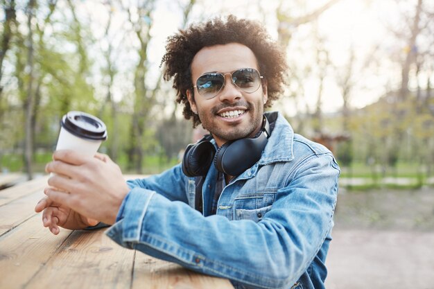 Außenporträt eines charmanten stilvollen dunkelhäutigen Mannes mit Afro-Frisur, der eine trendige Brille und einen Jeansmantel trägt, während er auf der Straße ist, sich auf den Tisch stützt und Kaffee trinkt