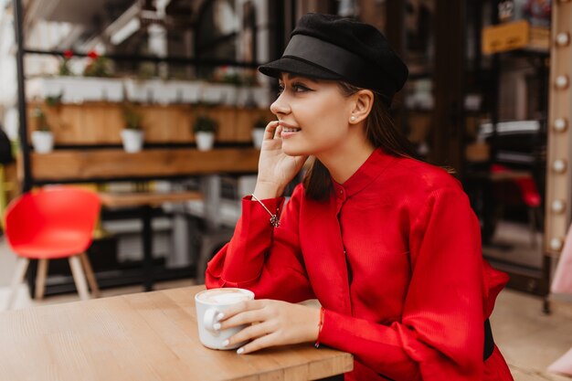 Außenporträt im Modellprofil mit üppigen Wimpern. Hübsches Mädchen trinkt Cappuccino und lächelt schüchtern