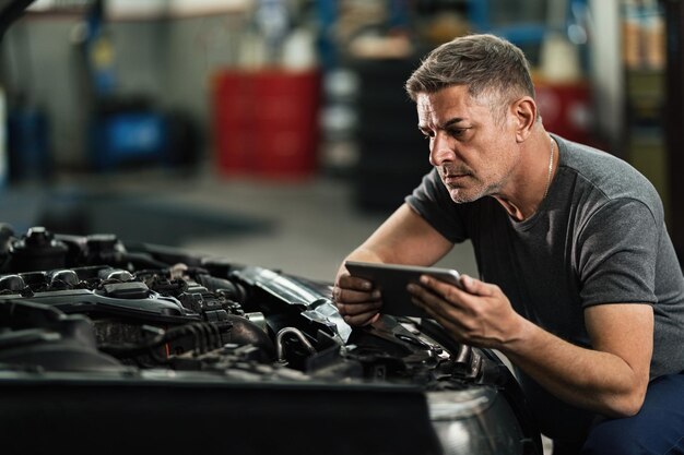 Automechaniker, der ein digitales Tablet verwendet, während er den Analysemotor in der Autowerkstatt repariert