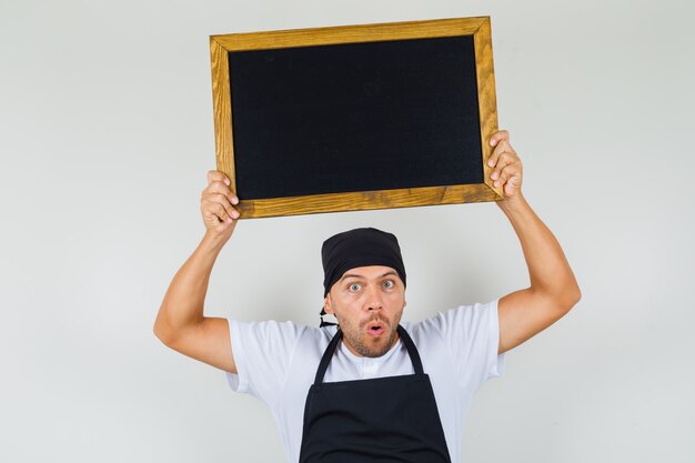 Kostenloses Foto bäcker mann hält tafel über den kopf in t-shirt, schürze und sieht überrascht aus