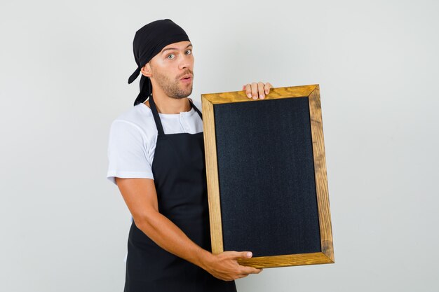 Bäcker Mann im T-Shirt, Schürze hält Tafel und sieht verwirrt aus