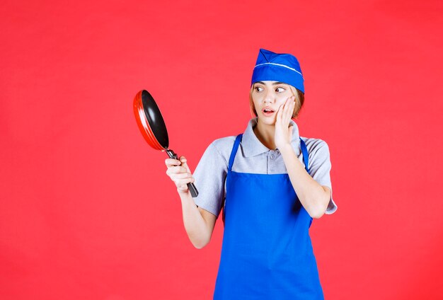 Bäckerin in blauer Schürze, die eine Tefal-Pfanne hält und verwirrt und nachdenklich aussieht
