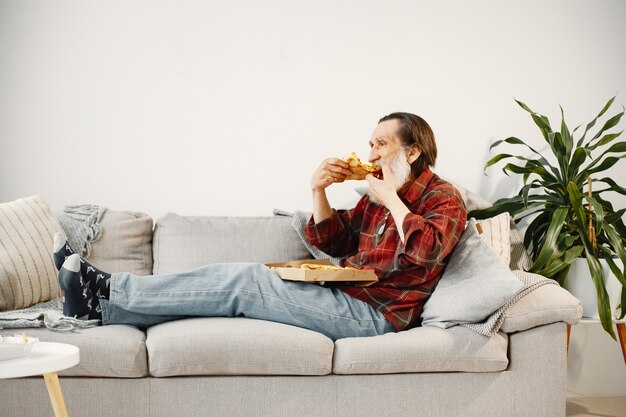 Bärtiger älterer Mann, der auf Couch liegt und Pizza isst. Fastfood.