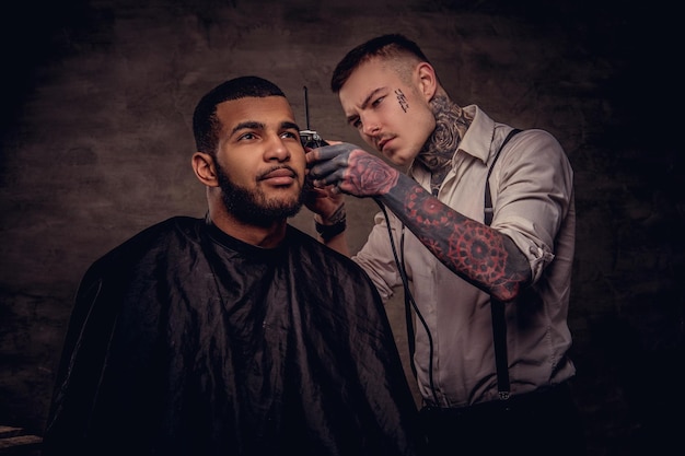 Bärtiger afroamerikanischer Hipster-Typ, der von einem altmodischen, tätowierten professionellen Friseur die Haare geschnitten bekommt, macht den Haarschnitt.