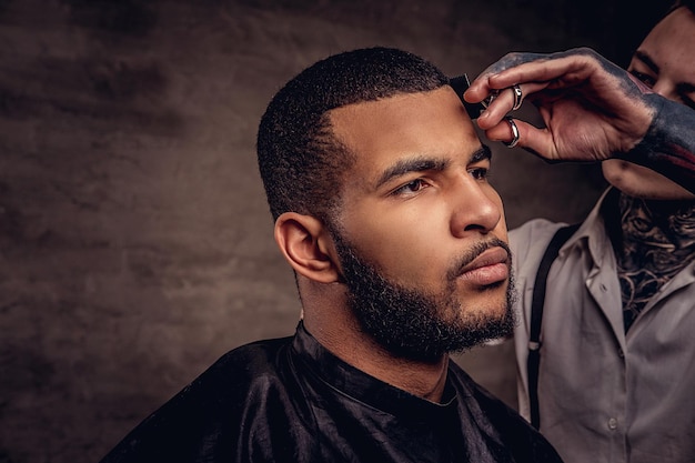 Bärtiger afroamerikanischer Hipster-Typ, der von einem altmodischen, tätowierten professionellen Friseur die Haare geschnitten bekommt, macht den Haarschnitt.