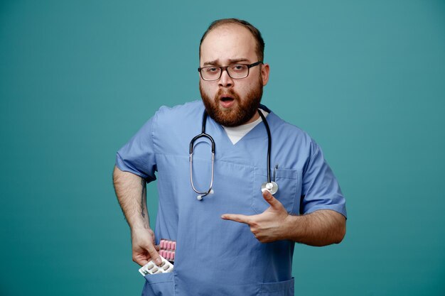 Bärtiger Arzt im blauen Mantel mit Stethoskop um den Hals, der eine Brille trägt, die Pillen aus der Tasche nimmt und verwirrt und überrascht über blauem Hintergrund steht