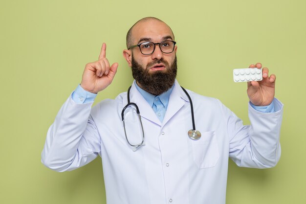 Bärtiger Arzt im weißen Kittel mit Stethoskop um den Hals, der eine Brille trägt und eine Blase mit Pillen hält, die mit den Zeigefingern nach oben zeigen und überrascht auf grünem Hintergrund stehen