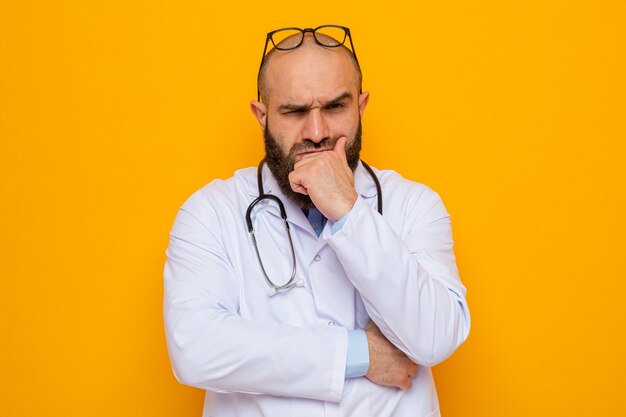 Bärtiger Arzt im weißen Kittel mit Stethoskop um den Hals mit Brille auf dem Kopf, der mit stirnrunzelndem Gesicht mit der Hand am Kinn schaut